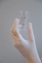 Load image into Gallery viewer, Hand with glove holding a chemistry baker with a house shaped gold ring
